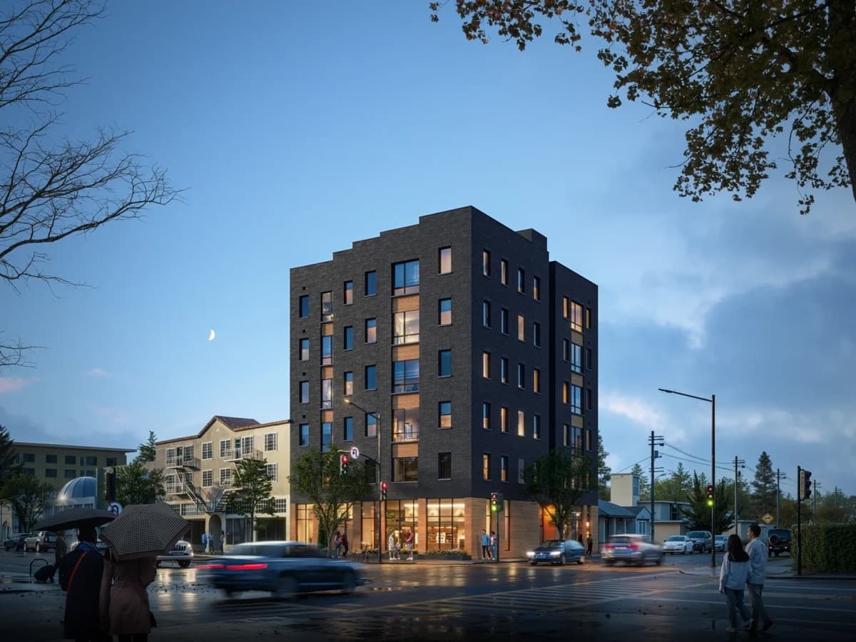Rendering of a seven-story dark brick apartment building with lit ground-floor shops on a rainy street corner at dusk